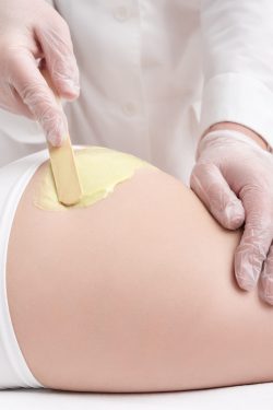 Unrecognizable beautician in gloves applying green hot wax on buttocks of slim woman using spatula. Waxing procedure, depilation process with hot wax in professional beauty salon. Part of photo series